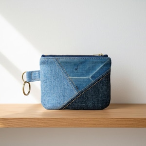 May include: A rectangular denim coin pouch featuring a patchwork design of light and dark blue fabric panels. The pouch includes a gold-toned metal zipper closure along the top edge and a matching denim side tab with a circular metal key ring attached.
