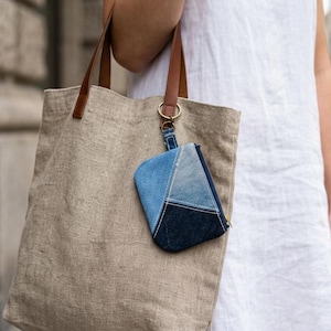May include: A beige linen tote bag with brown leather handles featuring a small, rectangular denim pouch attached to the handle by a brass ring. The pouch is constructed from patchwork denim fabric in varying shades of blue, from light to dark.