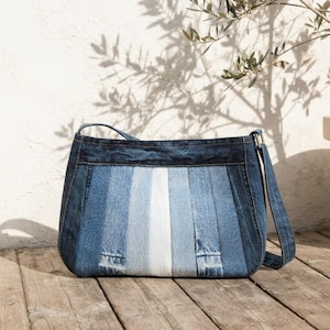 May include: A shoulder bag constructed from vertical panels of denim fabric in varying shades of blue, ranging from dark indigo at the edges to a light, faded white-blue center. The bag features a rectangular shape with rounded corners.