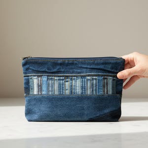 Peut inclure: Une pochette rectangulaire zippée fabriquée en tissu denim bleu foncé. L'avant présente une bande horizontale composée de bandes verticales de denim dans des nuances variées de bleu et bleu clair, avec des coutures visibles et une fermeture éclair.