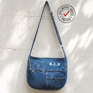 Op de afbeelding: Een blauwe denim schoudertas met een lange verstelbare riem, een rechthoekige Southpole-merkpatch en meerdere buitenzakken met riemlusdetails. De tas heeft een gebogen onderkant en is gemaakt van gerecyclede denimstof.