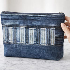 Könnte beinhalten: Eine rechteckige Denim-Tasche mit Reißverschluss. Die Tasche besteht aus blauem Denim mit einem helleren blau-weißen rechteckigen Design auf der Vorderseite. Die Tasche wird von einer Hand gehalten.