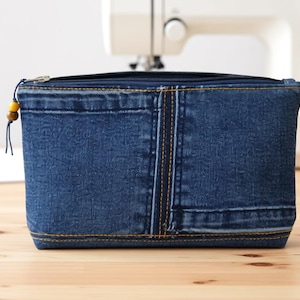Op de afbeelding: Een rechthoekig denim etui met ritssluiting. De etui is gemaakt van gerecycled spijkerbroekmateriaal, met zichtbare naden en stiksels. Een kleine houten kraal siert de ritstrekker. De achtergrond bevat een naaimachine.