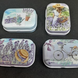 Op de afbeelding: Vier kleine rechthoekige metalen blikken met verschillende lavendel-thema ontwerpen. De blikken tonen een fiets, een vrouw met een mand vol lavendel, een gieter en een hoed.