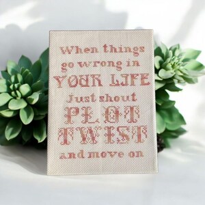 Puede incluir: Una pieza de arte mural rectangular de punto de cruz con el texto "When things go wrong in YOUR LIFE Just shout PLOT TWIST and move on" en hilo marrón rojizo. El fondo es de tela beige claro. Suculentas verdes enmarcan la obra de arte.