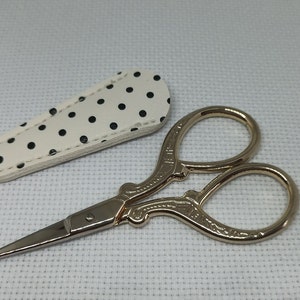 Könnte beinhalten: Goldfarbene dekorative Schere mit einer weißen Ledertasche mit schwarzen Punkten.