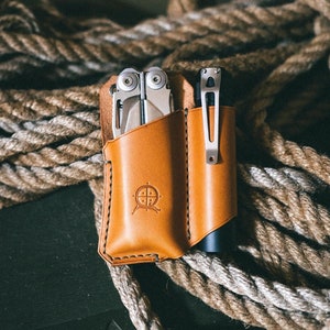 以下が含まれることがあります： コンパスロゴが付いた、茶色の革製のマルチツールと懐中電灯ホルダー。ホルダーはロープの山の上に置かれています。