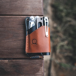 Może przedstawiać: Brązowa skórzana saszetka na narzędzia wielofunkcyjne z czarną podszewką i obszyciem. Saszetka ma małe logo z przodu i zawiera wielofunkcyjne narzędzie i małą latarkę.