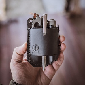 Puede incluir: Un organizador EDC de cuero negro con un bolígrafo, un cuchillo y otras herramientas. El organizador tiene un emblema decorativo y se sostiene en una mano. El bolígrafo es plateado y el cuchillo es gris.