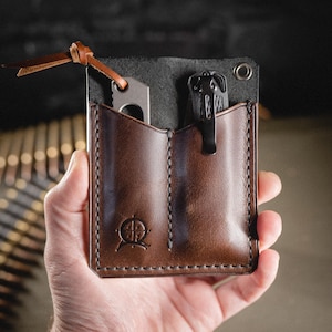 Könnte beinhalten: Ein brauner Leder-Taschenorganizer mit zwei Fächern, der ein kleines Messer und einen schwarzen Clip enthält. Der Organizer hat ein genähtes Design und ein Logo auf der Vorderseite. Ein Lederriemen ist oben befestigt.