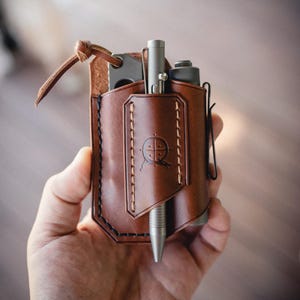 Op de afbeelding: Een bruin lederen multi-tool organizer met een pen, mes en andere gereedschappen. De organizer heeft een gestikt ontwerp en een logo op de voorkant. Een zwarte clip is aan de zijkant bevestigd.