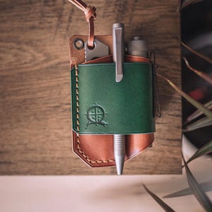 Puede incluir: Un organizador de bolsillo de cuero verde y marrón con un bolígrafo, un cuchillo y una linterna. El organizador tiene un diseño cosido y un lazo de cuero. El bolígrafo es plateado y el cuero está grabado con un diseño.