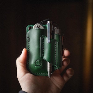 May include: A green leather EDC organizer with a pen, knife, and flashlight. The organizer has a stitched design and a logo on the front. The items are held in place by the organizer's pockets and loops.