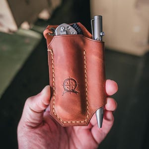 Peut inclure: Un étui de poche en cuir marron avec un motif cousu et un logo de boussole. L'étui contient un stylo argenté et un petit couteau de poche argenté.