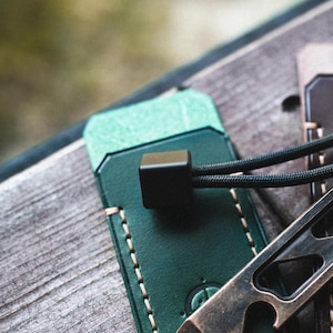 Peut inclure: Un étui en cuir vert foncé avec des coutures claires, doté d'une perle carrée noire attachée à un cordon noir. Un outil levier métallique à la finition vieillie avec une découpe décapsuleur est positionné à côté de l'étui en cuir.