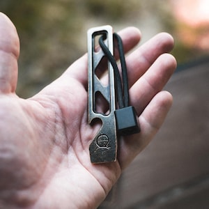 Puede incluir: Un abrebotellas rectangular de metal en tono bronce con una serie de recortes triangulares en el centro, un cordón negro con una cuenta cuadrada y un pequeño logotipo circular grabado cerca del borde inferior de la herramienta.