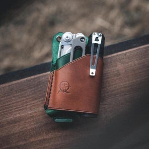 Könnte beinhalten: Ein brauner Lederhalter für Multitool und Taschenlampe mit grüner Innenausstattung. Der Halter hat ein geprägtes Kompasslogo auf der Vorderseite.
