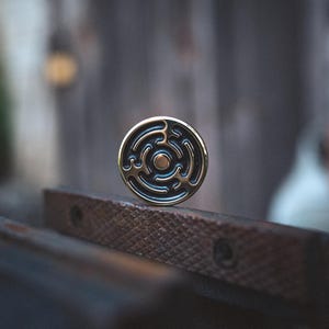Puede incluir: Un objeto metálico redondo, color latón, con un diseño de laberinto. El objeto tiene un centro oscuro y circular y un borde exterior dorado. Descansa sobre una superficie marrón texturizada sobre un fondo borroso.