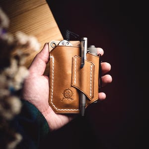 Könnte beinhalten: Ein brauner Leder-Taschenorganizer mit weißen Nähten, der einen Stift und ein kleines Messer enthält. Der Organizer hat ein geprägtes Emblem und eine Schlaufe für ein Schlüsselband. Der Stift ist silberfarben.
