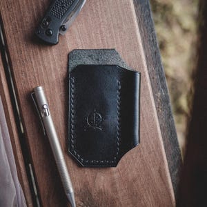 May include: A black leather wallet with a stitched border and a silver pen and a black folding knife on a wooden surface. The wallet has a debossed emblem. The knife is closed. The pen is silver.