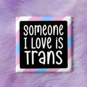 Puede incluir: Un botón cuadrado con un borde a rayas rosa, azul y blanco. El botón tiene un fondo negro con el texto "someone I love is trans" en blanco.