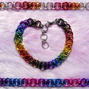 May include: A heart-shaped bracelet and two chain bracelets made of colourful metal rings. The heart-shaped bracelet features a rainbow of colours, while the other two bracelets display a gradient of colours, including silver, red, and purple. The jewellery is displayed on a pink surface.