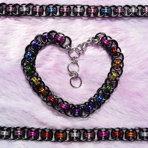 May include: A heart-shaped black chainmail bracelet with a silver clasp, featuring colourful rings in red, orange, yellow, green, blue, and purple. Two matching chainmail necklaces are also visible.