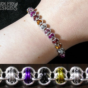 Könnte beinhalten: Ein silbernes Kettenarmband mit abwechselnd gelben, orangefarbenen, roten, violetten und schwarzen Ringen. Das Armband ist aus Metall und hat ein geflochtenes Design. "Faerie Fire Design" ist im Hintergrund gedruckt.