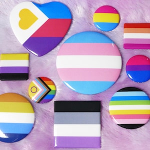 Może przedstawiać: Zbiór dziesięciu okrągłych i kwadratowych pinów z różnymi wzorami flag LGBT. Piny są w kolorach takich jak żółty, niebieski, różowy, biały, czarny, fioletowy, zielony i pomarańczowy.