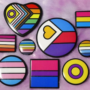 May include: A set of nine colorful enamel pins featuring various pride flags, including the transgender, lesbian, pansexual, and bisexual flags. The pins are all round or heart-shaped and have a black outline.