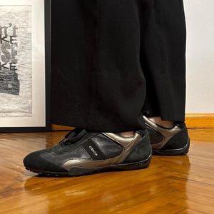 Puede incluir: Zapatillas deportivas negras y doradas metalizadas con la marca "Geox" visible en el lateral. Las zapatillas se llevan con pantalones negros. En el fondo, una impresión enmarcada con las palabras "Don't Make Sense Make".