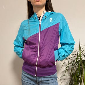 May include: A teal and purple Scott windbreaker jacket with a white zip and a hood. The jacket features a diagonal colour block design and a white logo on the chest. The model is wearing blue jeans.