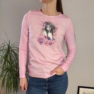 以下が含まれることがあります： 花柄の王冠と花をあしらった女の子の水彩画が特徴の、明るいピンク色の長袖シャツ。 シャツは柔らかい素材でできており、クルーネックです。