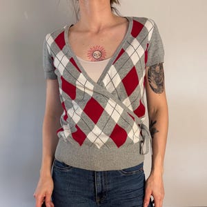 May include: A short-sleeved wrap top featuring a grey, red, and white argyle diamond pattern. The knit garment has a V-neckline, a ribbed hem, and a side tie closure. It is worn over a light-colored camisole, paired with blue denim jeans.