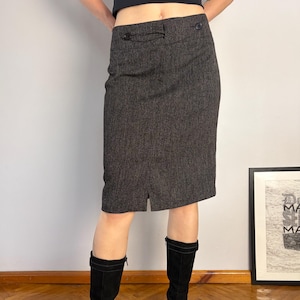 May include: A charcoal gray knee-length skirt featuring a textured fabric, a fitted waistband with two decorative buttons, and a small vertical slit at the center front hem. The skirt is paired with black mid-calf boots and a black sleeveless top.