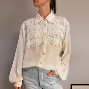 May include: A long-sleeved white button-down shirt featuring a pointed collar and three horizontal rows of intricate white lace trim across the chest. The shirt is fastened with round, gold-rimmed buttons and has a loose, flowing fit.