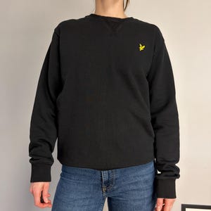 Puede incluir: Una sudadera negra de manga larga con cuello redondo, que presenta un cuello acanalado con un detalle de costura en forma de V en el escote y un peque&ntilde;o logotipo de p&aacute;jaro bordado en amarillo en el pecho izquierdo, junto con vaqueros azules.