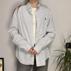 May include: A white button-down shirt with a blue and black checkered pattern. The shirt has long sleeves and a small black logo on the chest. The shirt is paired with gray jeans.