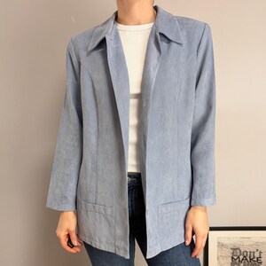 May include: A light blue blazer with a collared neckline, long sleeves, and two front pockets. The jacket features a smooth, matte texture and a button-front closure. It is worn over a white ribbed shirt and dark blue denim jeans.