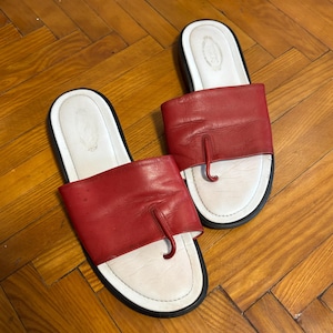 May include: A pair of red leather sandals with white soles and black trim. The sandals have a thong-style design and are displayed on a wooden floor.