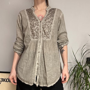 Puede incluir: Camisa de manga larga marrón claro con un panel de lentejuelas en la parte delantera. La camisa tiene cuello mandarín y cierre de botones. Las mangas están enrolladas. La camisa se combina con pantalones negros.