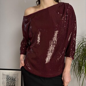 May include: A burgundy sequined top with an off-the-shoulder design. The top has three-quarter sleeves and a loose fit. The model is wearing black pants. A framed print with the text "Don't Make Sense Make" is in the background.