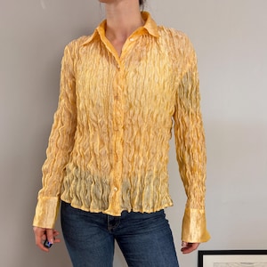 Pode incluir: Uma camisa de manga comprida com botões num tom amarelo vibrante, apresentando um tecido texturizado e amarrotado em toda a peça. A camisa tem um colarinho clássico, uma carcela de botões frontal e punhos largos e lisos, combinada com jeans azuis.