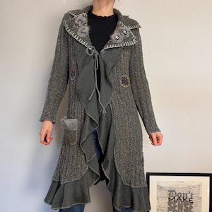 May include: A long, dark gray, ribbed knit cardigan featuring a ruffled front edge, a tie closure, and decorative circular patches with faux fur accents. The garment includes a patch pocket with faux fur trim and white contrast stitching along the collar.