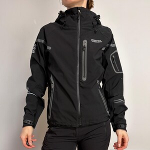 May include: A black technical jacket featuring a hood with adjustable drawstrings, a full-length front zipper, and multiple zippered pockets. The jacket includes grey reflective accents on the sleeves and chest, with the Nordblanc logo printed on the upper right chest area.