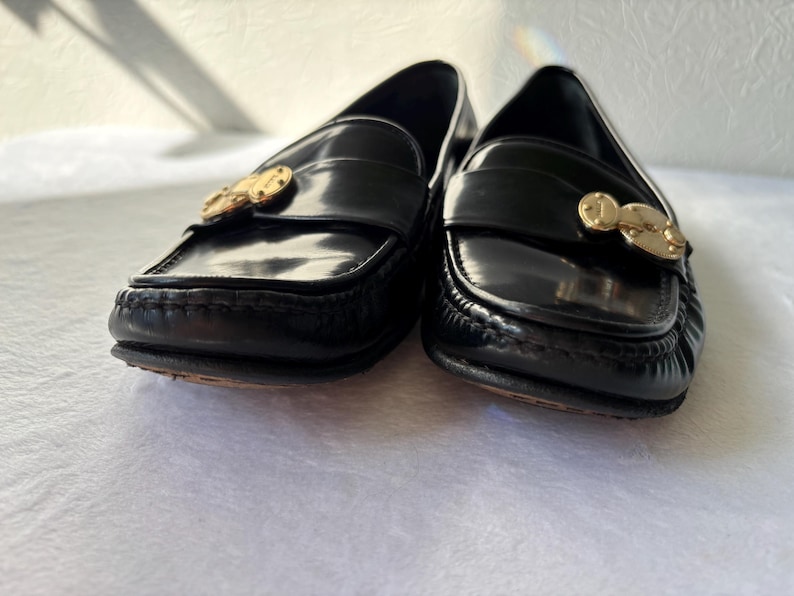 Może przedstawiać: Para czarnych sk&oacute;rzanych mokasyn&oacute;w ze złotymi detalami. Buty mają klasyczny design z zaokrąglonym noskiem i ozdobną klamrą. Podeszwy są w kolorze ciemnobrązowym.