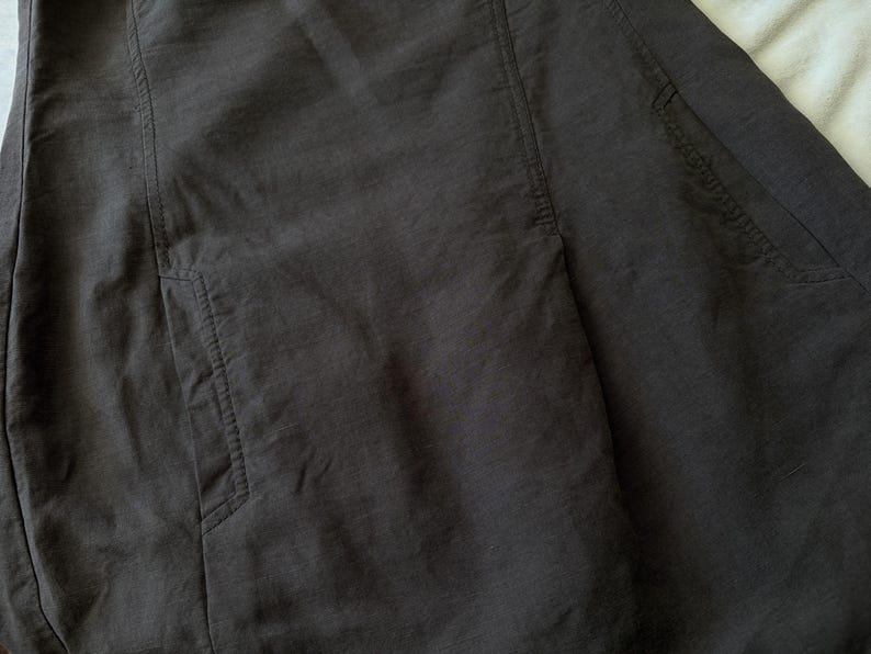May include: A dark charcoal gray garment featuring vertical seam detailing and two visible side pockets. The fabric has a subtle, slightly textured weave with visible stitching along the pocket openings and the vertical panel seams.