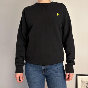 Puede incluir: Una sudadera negra de manga larga con cuello redondo y un detalle de costura en forma de V en el cuello. La prenda presenta un peque&ntilde;o logotipo de &aacute;guila bordado en amarillo en el pecho izquierdo y pu&ntilde;os acanalados en mangas y bajo.