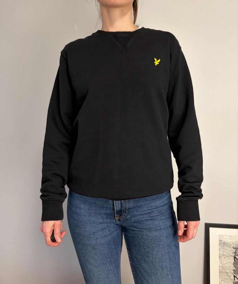 Puede incluir: Una sudadera negra de manga larga con cuello redondo y un detalle de costura en forma de V en el cuello. La prenda presenta un peque&ntilde;o logotipo de &aacute;guila bordado en amarillo en el pecho izquierdo y pu&ntilde;os acanalados en mangas y bajo.
