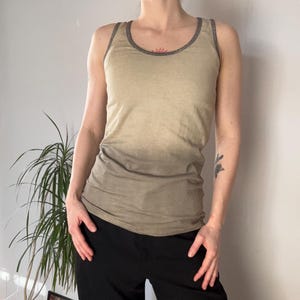Könnte beinhalten: Ein beiges Tanktop mit grauem Besatz am Ausschnitt und an den Armausschnitten. Es wird mit einer schwarzen Hose kombiniert. Das Tanktop ist ärmellos und hat einen Rundhalsausschnitt. Der Stoff scheint ein weiches, leichtes Material zu sein.